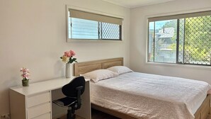 Une chambre à coucher comprenant un lit, un bureau avec une chaise, un ventilateur de plafond et deux fenêtres.