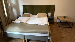 Uma cama bem arrumada com cabeceira verde, duas mesas de cabeceira com abajures e uma pequena mesa de madeira com uma planta.