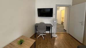 Quarto pequeno e moderno com televisão de tela plana na parede, uma escrivaninha com cadeira, um ventilador e uma mesinha com planta.