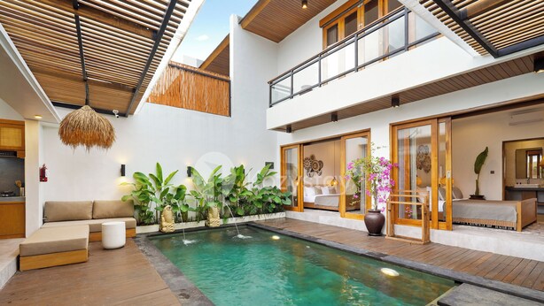 A modern indoor pool area with a wooden deck, a seating area with cushions, and a variety of plants.