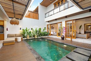 A modern indoor pool area with a wooden deck, a seating area with cushions, and a variety of plants. - Kastara Villa by Kozystay - Canggu (Canggu)