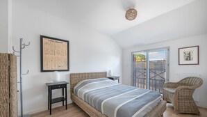 Un dormitorio con una cama de mimbre, una ilustración botánica enmarcada, una silla de mimbre y vistas a través de un ventanal.