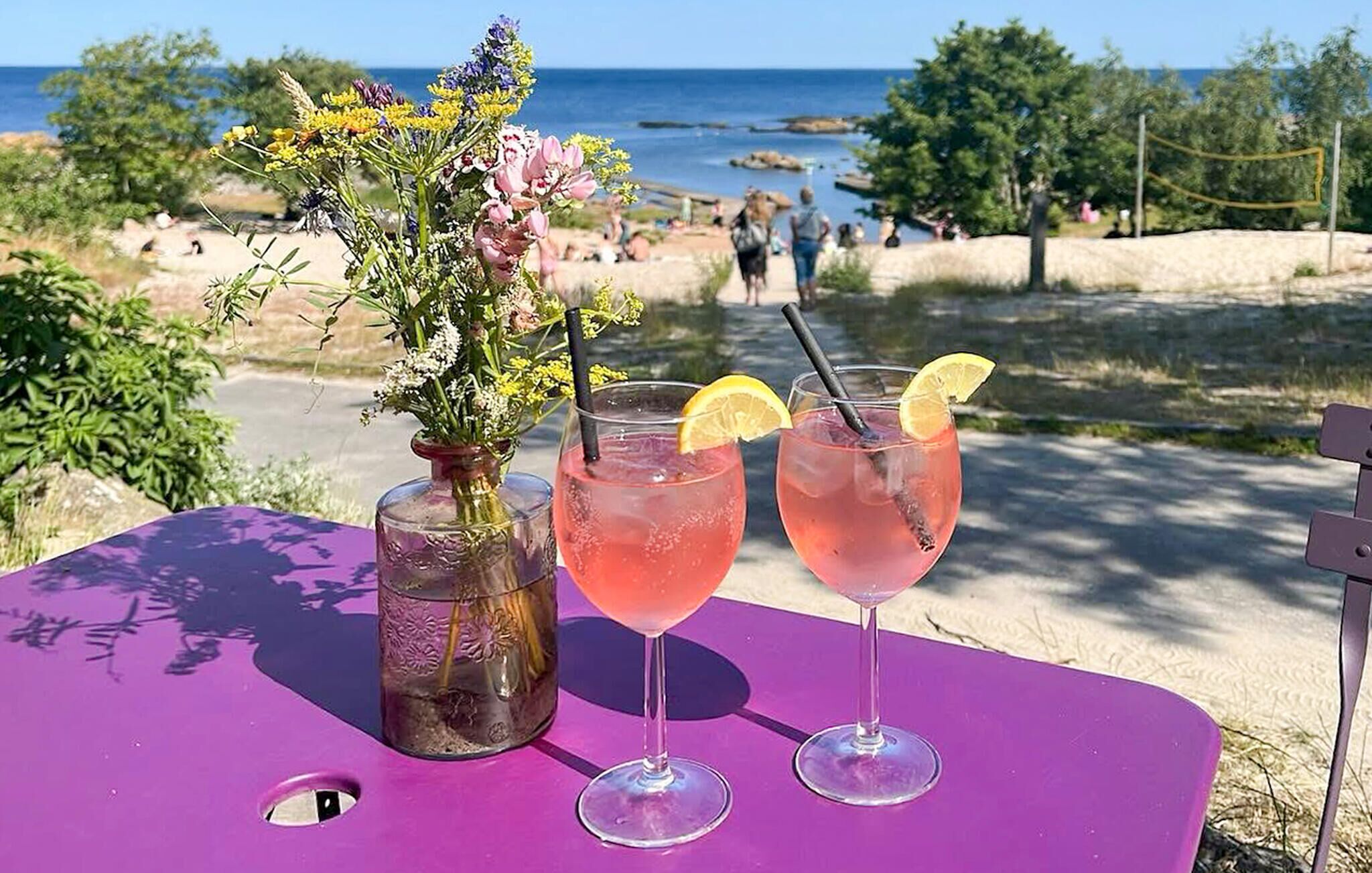 A table with two glasses of pink drink, a vase with flowers, and a view of the beach and ocean.