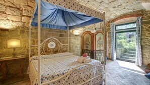Une chambre à coucher avec un lit à baldaquin, des murs en pierre, une table de chevet en bois, une lampe et une vue sur la verdure par la fenêtre.