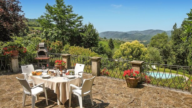 Un espace extérieur aménagé pour les repas, avec un sol en pierre, des chaises blanches, une table dressée pour un repas, un barbecue en pierre et une vue sur une piscine et des montagnes.