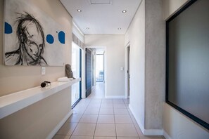 Un couloir moderne au design minimaliste, avec un sol carrelé blanc, une œuvre d’art accrochée au mur et une étagère intégrée sur laquelle sont posés une statue et une serviette.