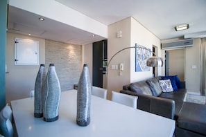 Un salon moderne avec une table blanche, deux vases décoratifs, un canapé avec des coussins, un tableau et un luminaire au plafond.