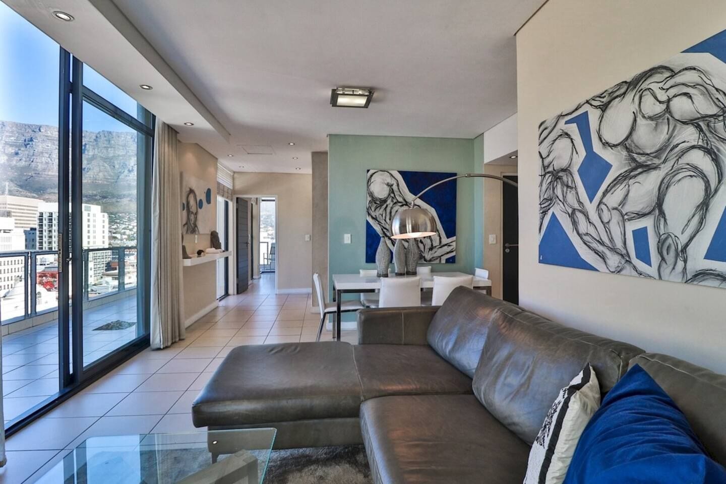 A modern living room with a leather sofa, a dining area, and large windows offering a city view.