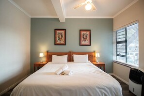 Una camera d'albergo con un letto, due abat-jour, una ventola a soffitto e due quadri appesi al muro.