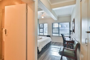 Una camera d'albergo con un letto, una scrivania, una sedia, una ventola a soffitto e due finestre con tende.
