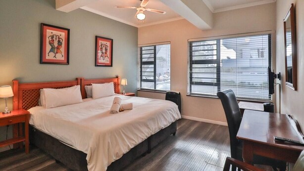 A hotel room with a bed, a desk, a chair, a ceiling fan, and two windows with blinds. - Spring Tide Inn - Standard king room 3 (Cape Town)
