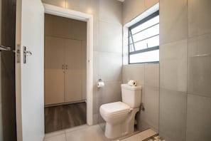 A bathroom with a toilet, a roll of toilet paper, a window, and a door leading to a room with cabinets.