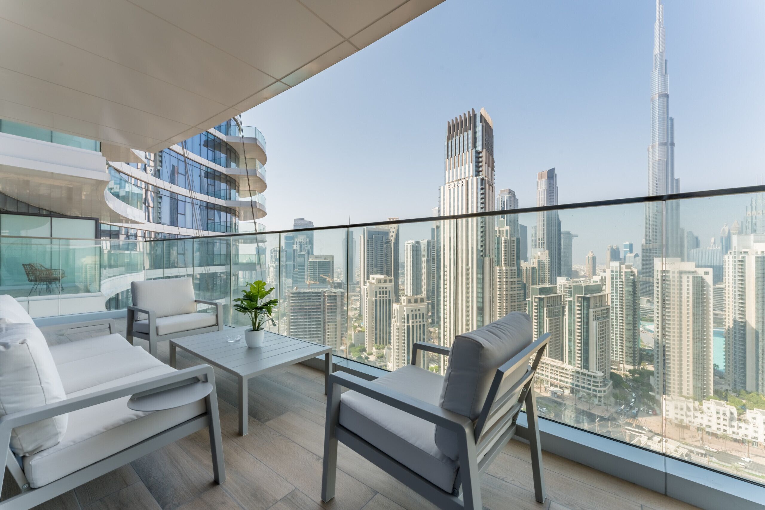 A modern balcony with a glass railing, white furniture, and a view of a cityscape including a tall skyscraper.