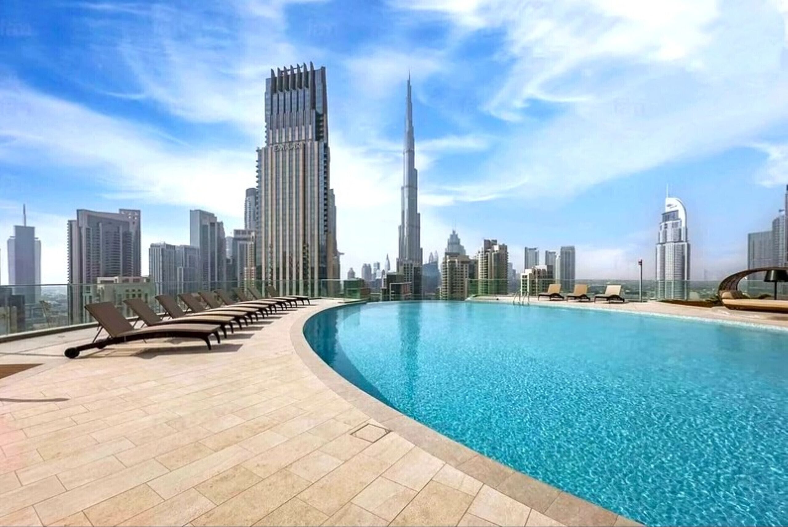 A rooftop pool with lounge chairs, a city skyline in the background, and modern high-rise buildings.