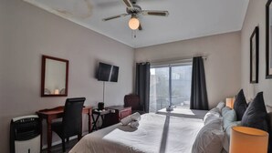 A hotel room with a bed, desk, chair, TV, and a large window with curtains. - Spring Tide Inn - Std family room 7 with balcony (Cape Town)