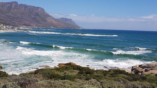 A coastal landscape with rocky terrain, waves, and a distant mountain range. - Spring Tide Inn - Std King Room 8 with Balcony (Cape Town)