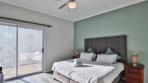 A bedroom with a large bed, a ceiling fan, a nightstand, and a sliding glass door leading to an outdoor area. - Spring Tide Inn - Std King Room 8 with Balcony (Cape Town)