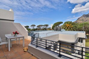 A rooftop terrace with a white table and chairs, a flower vase, and a view of trees and buildings.