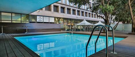 An indoor swimming pool with a wooden deck, a lifeguard stand, and a building with large windows in the background.