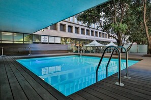 Una piscina interior con plataforma de madera, un puesto de socorrista y un edificio con grandes ventanales al fondo.