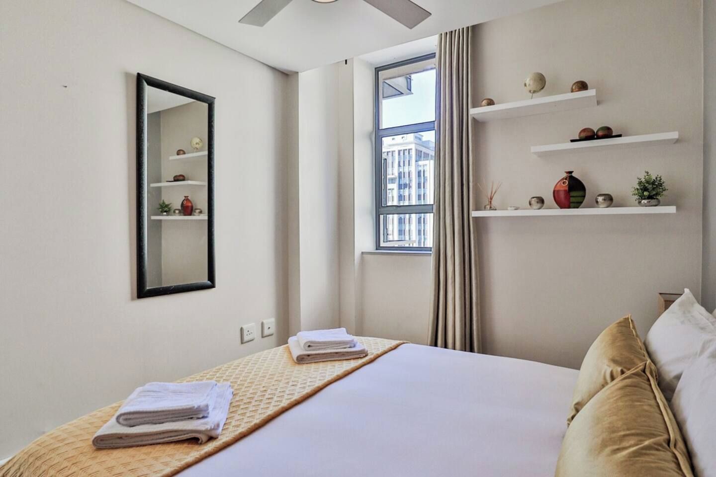 A hotel room with a bed, a mirror, a window with curtains, and shelves with decorative items.