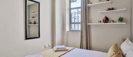 A hotel room with a bed, a mirror, a window with curtains, and shelves with decorative items.