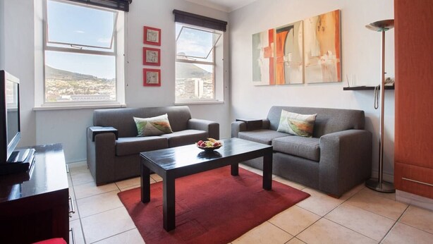 A living room with two sofas, a coffee table, and a TV. There are windows with blinds, a painting on the wall, and a floor lamp. - Piazza on Church 906 by Ctha (Cape Town)