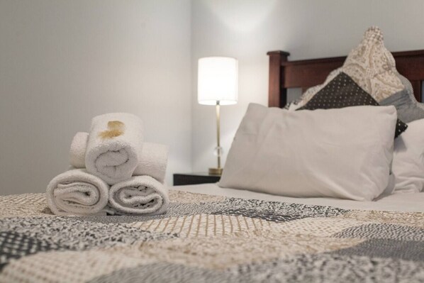A neatly made bed with rolled white towels, a patterned throw, and a bedside lamp.