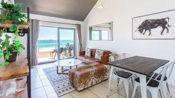 Un vaste espace de vie avec une grande fenêtre donnant sur la plage, un coin salon confortable et une table à manger avec des chaises.