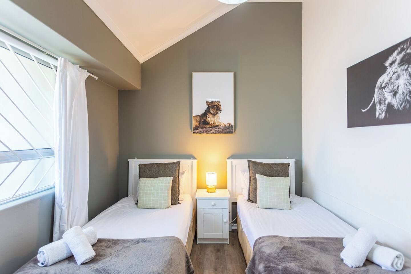 A room with two beds, a window with curtains, a nightstand with a lamp, and a painting on the wall.