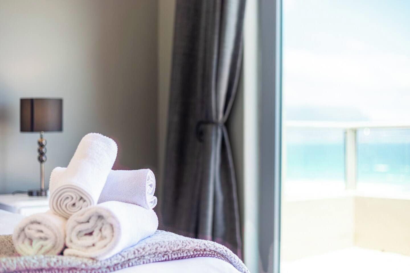 A neatly made bed with rolled white towels, a lamp, and a view of the sea through a window with curtains.
