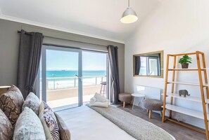 A room with a large sliding glass door leading to a balcony with a view of the sea. There is a bed with a white headboard, a small round table, and a wooden ladder with shelves.