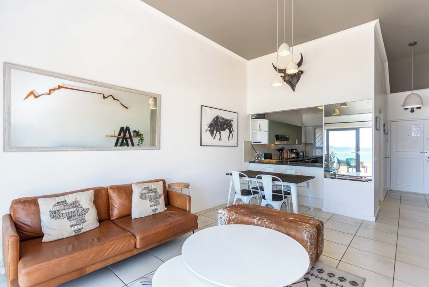 Una sala de estar moderna con un sofá de cuero marrón, una mesa redonda blanca y una cocina visible a través de una puerta abierta.