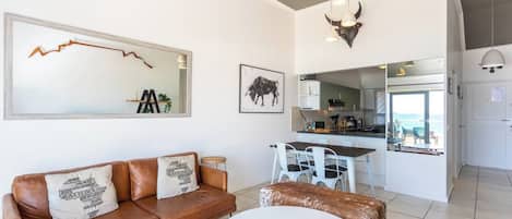 Una sala de estar moderna con un sofá de cuero marrón, una mesa redonda blanca y una cocina visible a través de una puerta abierta.