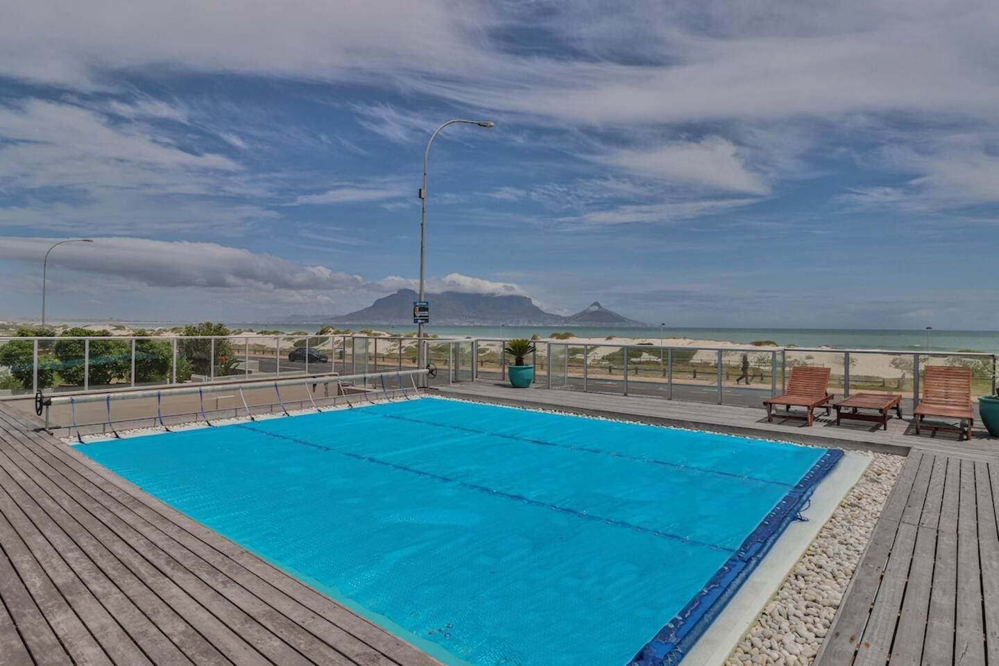Una piscina con terraza de madera, sillas de descanso y vistas a la playa y las montañas.
