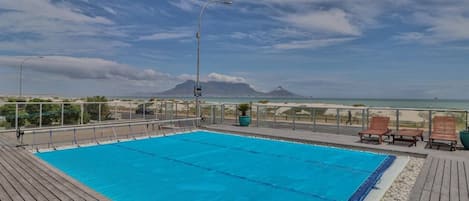 Una piscina con terraza de madera, sillas de descanso y vistas a la playa y las montañas.