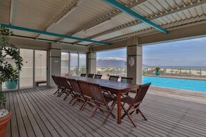 Una terrazza esterna con tavolo e sedie in legno, con vista su una piscina e una spiaggia.