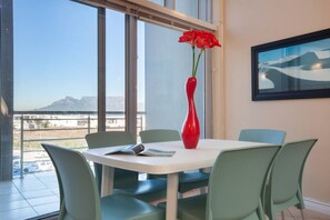 A dining area with a table, chairs, a red vase with flowers, and a painting on the wall.