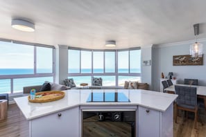 A modern kitchen with a central island, large windows, and a view of the sea.