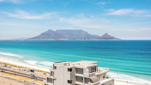 A coastal cityscape with a prominent mountain in the background, a beach with a road and vehicles in the foreground, and a modern building with balconies overlooking the sea. - Ocean View A1101 by Ctha (Cape Town)