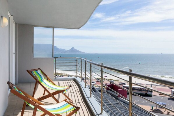 A balcony with a view of the sea and a mountain, two striped lounge chairs, and a bus in the background.