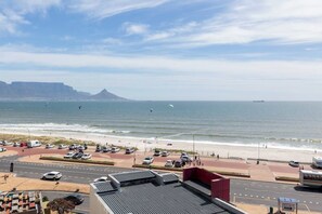 A coastal road with a beach and ocean view, vehicles parked along the roadside, and a distant mountain range.