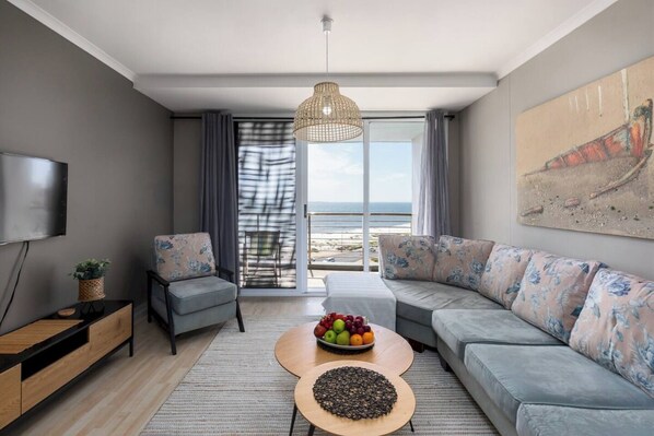 A modern living room with a sofa, a coffee table with fruit, a TV, and a view of the beach.