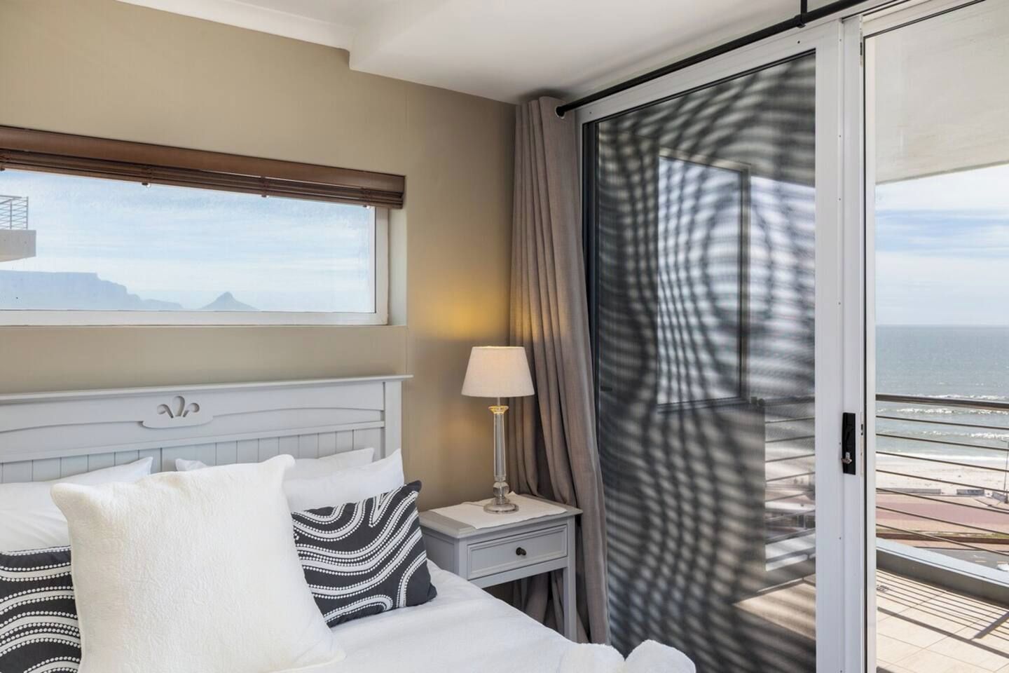 A hotel room with a bed, a nightstand, a lamp, a window with a view of the sea, and a sliding door to a balcony.