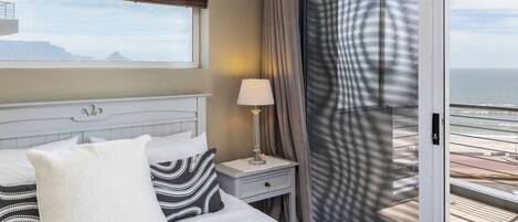 A hotel room with a bed, a nightstand, a lamp, a window with a view of the sea, and a sliding door to a balcony.