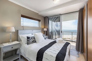 Una camera d'albergo con un letto, comodini, una finestra vista spiaggia e un balcone con tende.