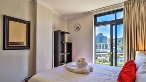 A hotel room with a bed, a red pillow, a clock, and a window with a city view. - Quayside 1206 by Ctha (Cape Town)