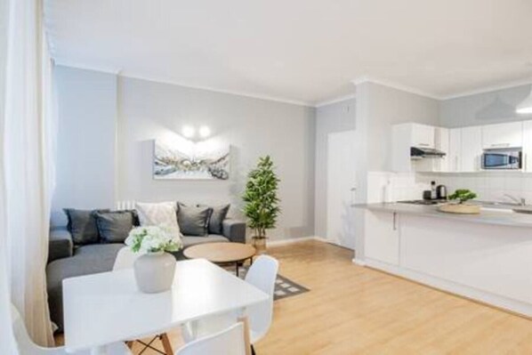 A modern apartment with a dining area, a living room with a sofa, a kitchen with white cabinets, and a potted plant.