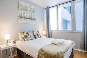 A hotel room with a bed, two bedside lamps, a window with curtains, and a painting on the wall.