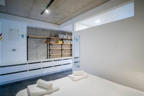 A minimalist room with a bed, a bench, and a wall-mounted shelf with boxes.